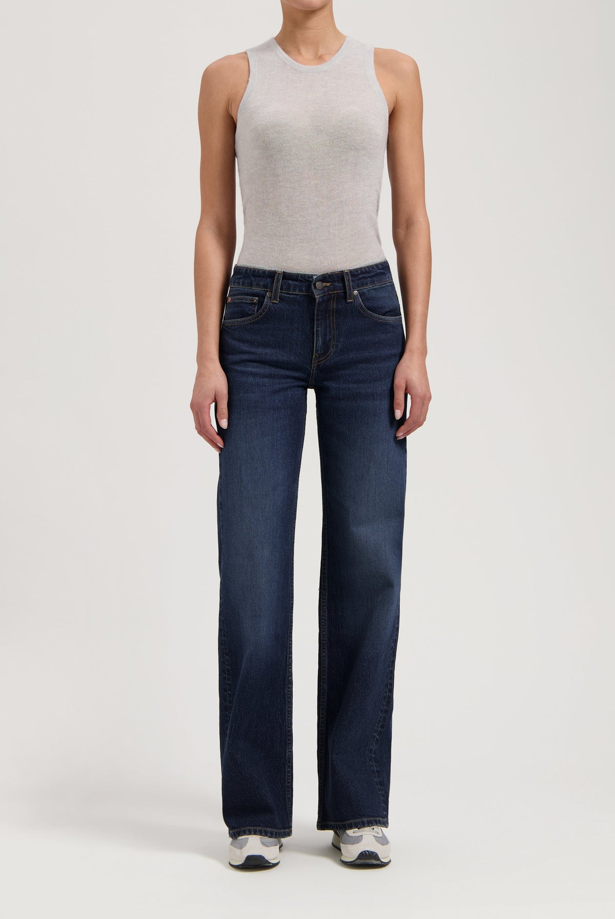 Woman wearing a light gray sleeveless top paired with dark blue, wide-leg jeans and white sneakers, standing in studio.