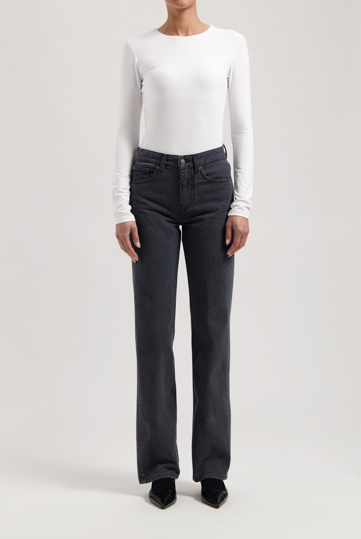 Model wearing high-waisted, black denim jeans paired with a white long-sleeve fitted top and black pointed shoes.