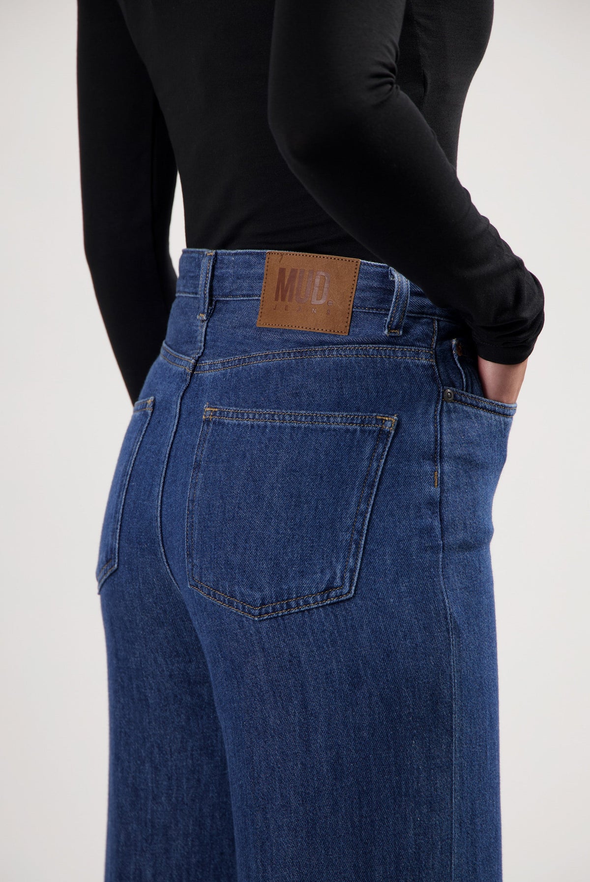 Person wearing blue MUD Jeans, showcasing sustainable high-waisted denim with a leather patch, paired with a black top.