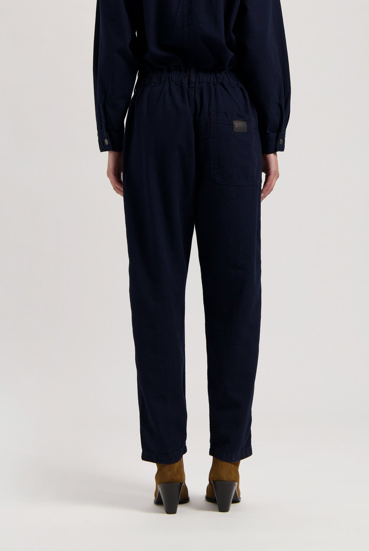Navy blue cotton jumpsuit with elastic waistband and buttoned cuffs, worn with brown heeled boots viewed from the back.