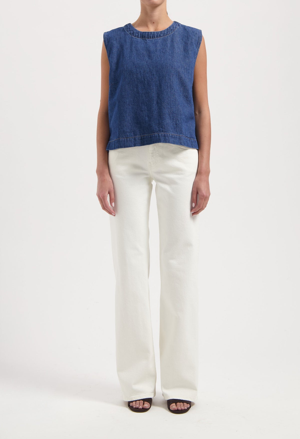 Woman wearing a sleeveless blue denim top paired with high-waisted white wide-leg jeans and black open-toe sandals.
